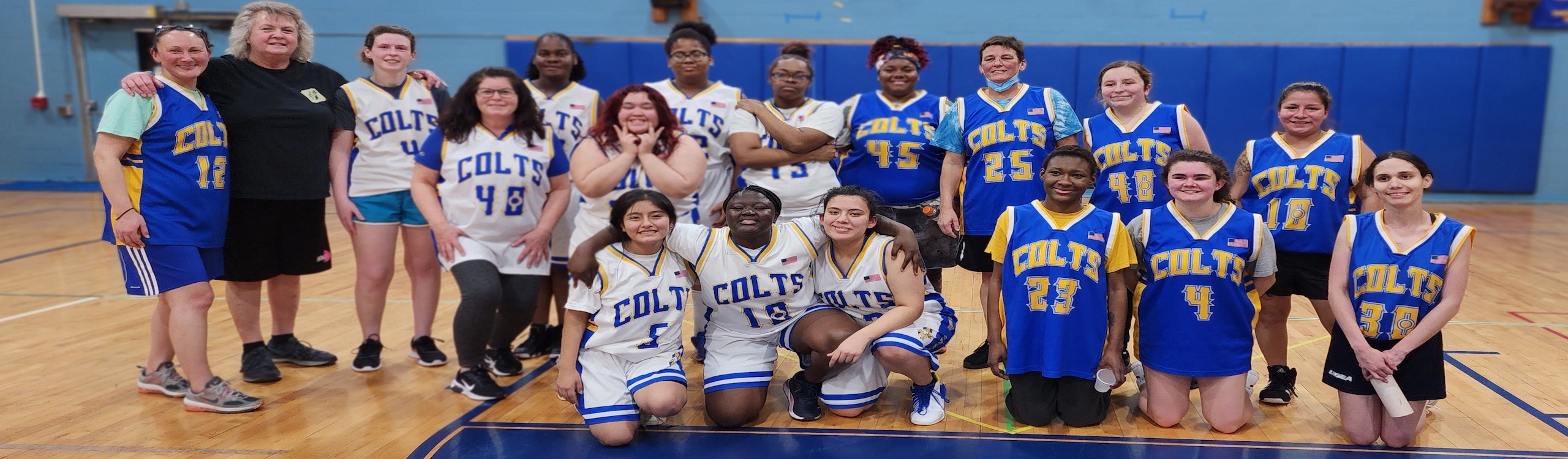 Group picture at foul line of women's alumni game participants. Students wearing white uniforms and alumni wearing blue uniforms.